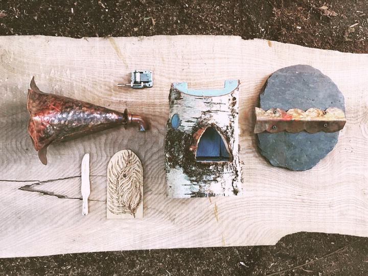 a photo of various elements - copper tube, carved wooden feather, bird's house and a piece of slate, all laid out nicely on a piece of thick wood
