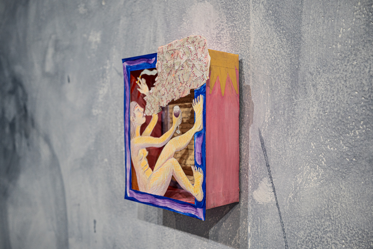 a small yellow box with a blue frame hanging on the grey wall showing a cut-out drawing of a girl sitting and drinking wine and smoking a cigarette. the smoke she is blowing out is made of paper mache and glitter. There re words written inside.