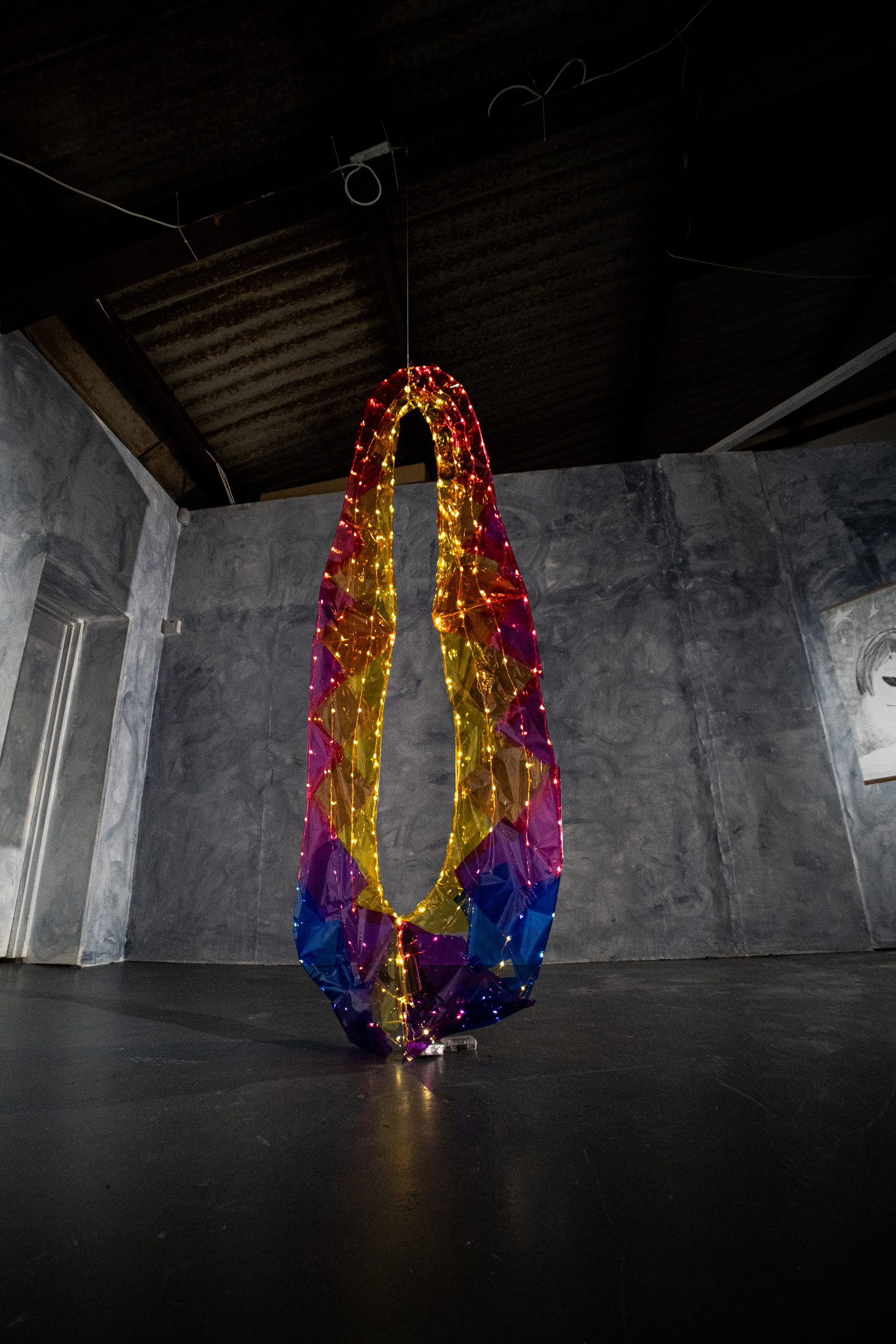 a photograph of a sculpture hanging from the ceiling of a building. it has a circular shape - like a portal and is multicoloured, rainbow coloured with fairy lights shining on it.