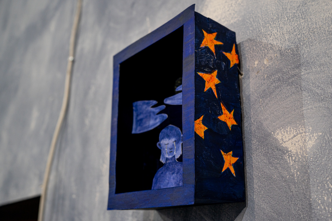 a blue box with orange stars on its walls is hanging on a grey wall. inside the box there is a cut-out drawing of a girl painted in blue with two glitter lines on her cheeks - she is crying. there are blue clouds above her head. everything is blue