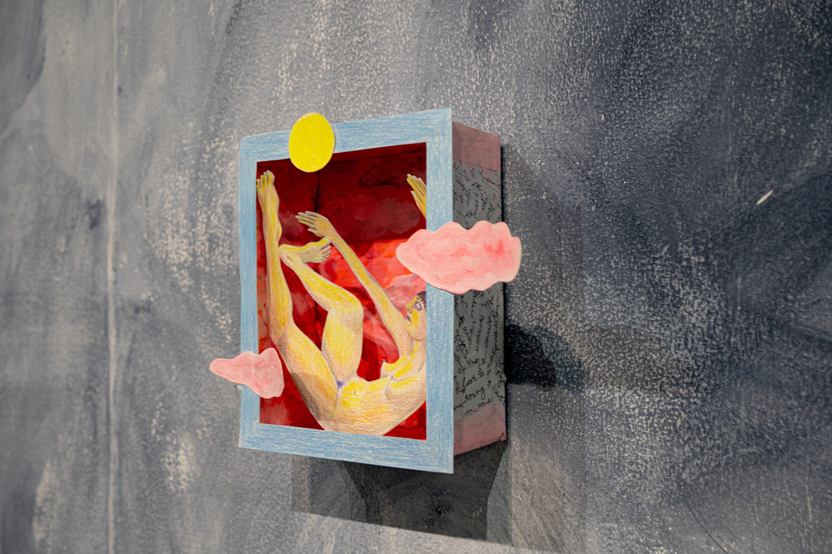 there is a small box hanging on a grey wall. Inside there is a cut-out drawing of a girl sitting in a box with a cut-out yellow sun above her and two pink cut-out clouds around her. there is writing around the outside walls of the box
